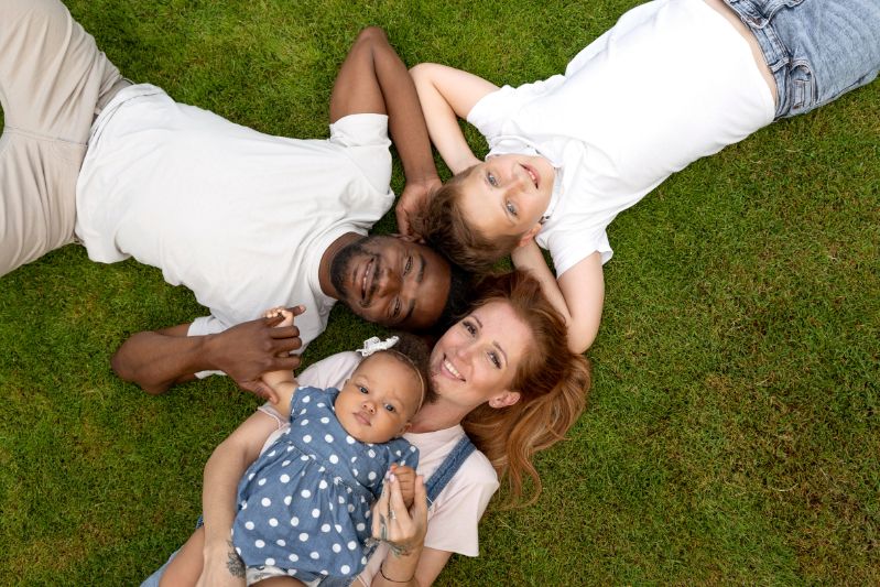 A family of four lying on green grass with their heads together in a circle, smiling up at the camera. A father with dark skin and short hair and a mother with long red hair are holding hands with their two children—a young boy in a white shirt and a baby girl in a blue polka-dot dress.