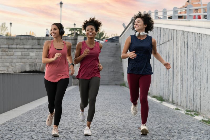 Three women jogging together on a cobblestone path outdoors at sunset, smiling and wearing athletic clothing and headphones, with a stone wall and street lamps in the background.